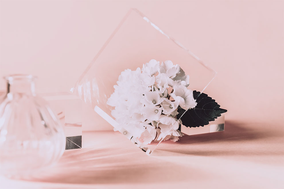 um bloco quadrado de acrílico com um pequeno buquê de flor branca ao fundo em cima de uma mesa rosa claro