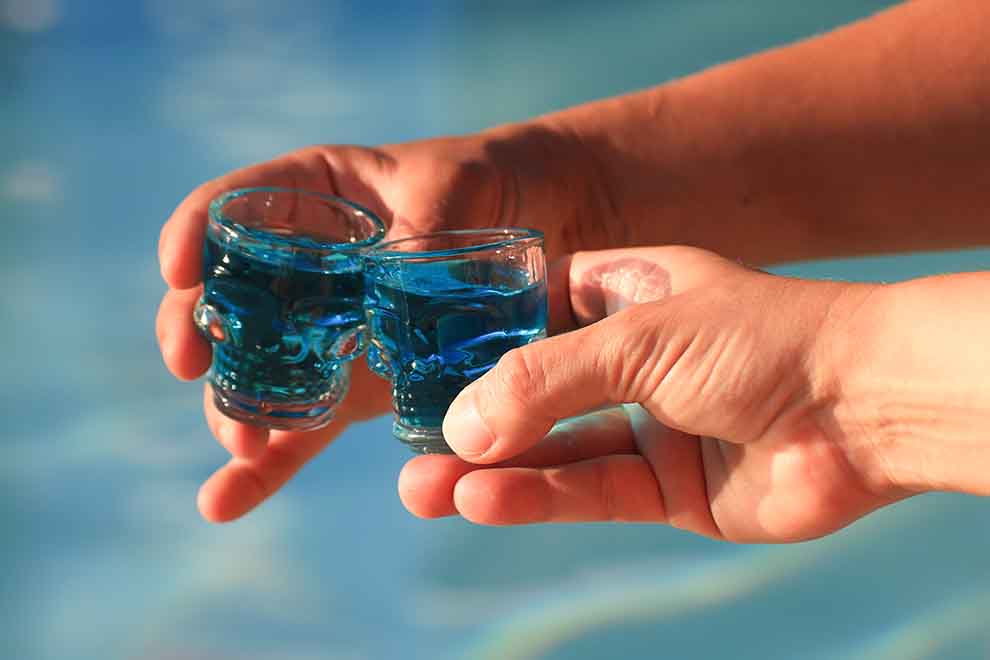 duas pessoas segurando copos de shot em forma de caveira com uma bebida azulada em uma praia