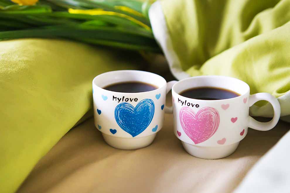 duas xicaras de casal personalizados com café dentro em cima de uma sofá bege em uma sala em um dia ensolarado