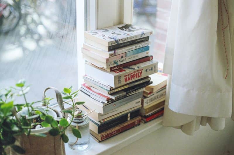 vários livros empilhados perto de uma janela e plantas e cortinas ao lado