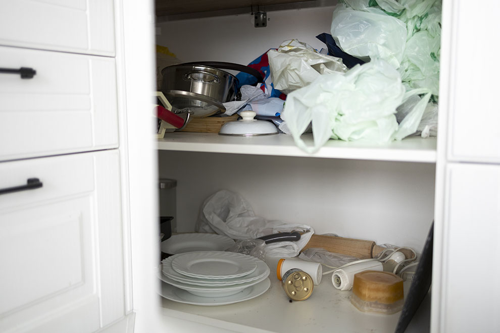 Armário de cozinha desorganizado e sujo, com panelas, sacolas de mercado e utensílios 