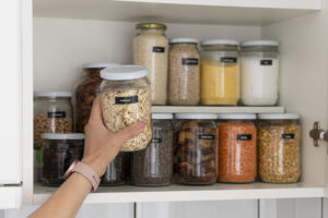 Interior de uma armário de cozinha com diversos potes com temperos, com uma mão feminina colocando um pote com aveia no armário