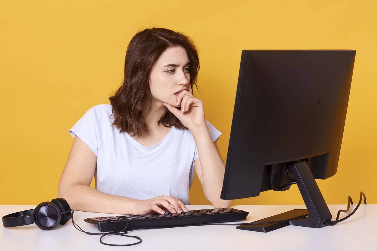 Modelo feminina, sentada em uma mesa com computador, escolhendo a melhor opção em seu computador, em um fundo amarelo. 