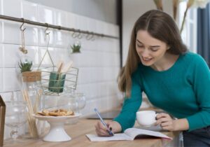 Moça escrevendo em caderno com uma xicara na mão