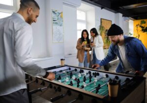 dois amigos jogando pebolim e duas amigas no fundo mexendo no celular