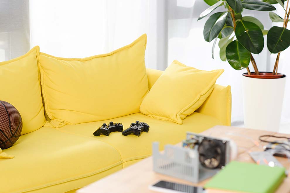 Sofá amarelo com controle de videogame e planta decorativa.