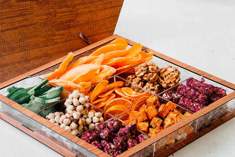 um recipiente de acrílico e madeira com diversas frutas secas dentro em um fundo branco