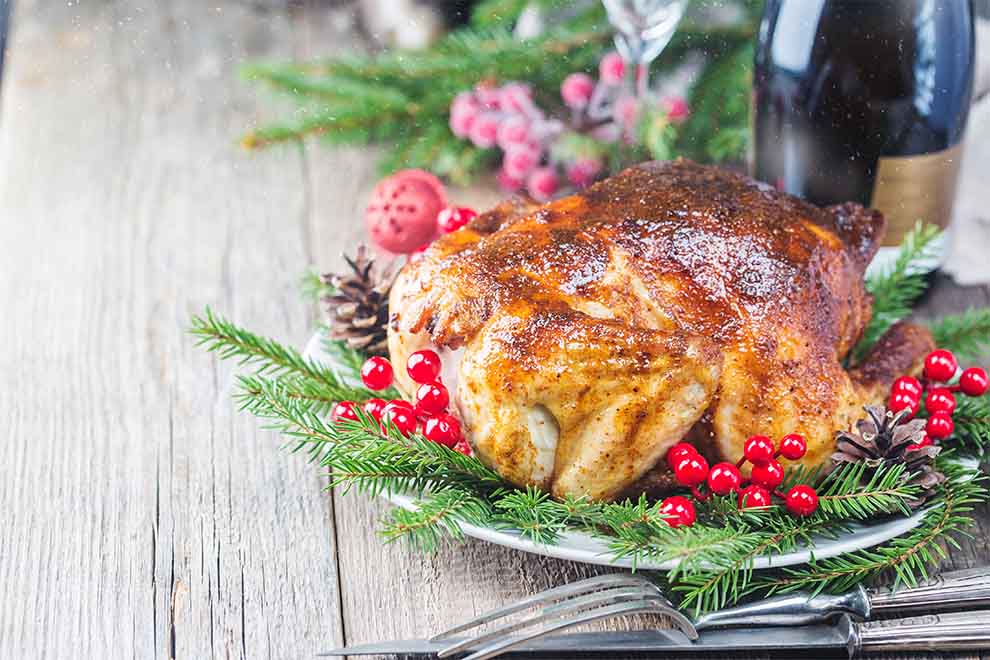 um peru de natal decorado com ramos de alecrim, pinha e pequenas frutas dentro de uma travessa branca de acrílico e talheres em cima de uma mesa de madeira 
