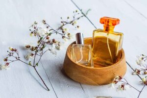 Imagem de um suporte para perfume redondo e de madeira, com dois perfumes sob ele, com dois ramos de flores ao lado, em cima de uma mesa de madeira cinza