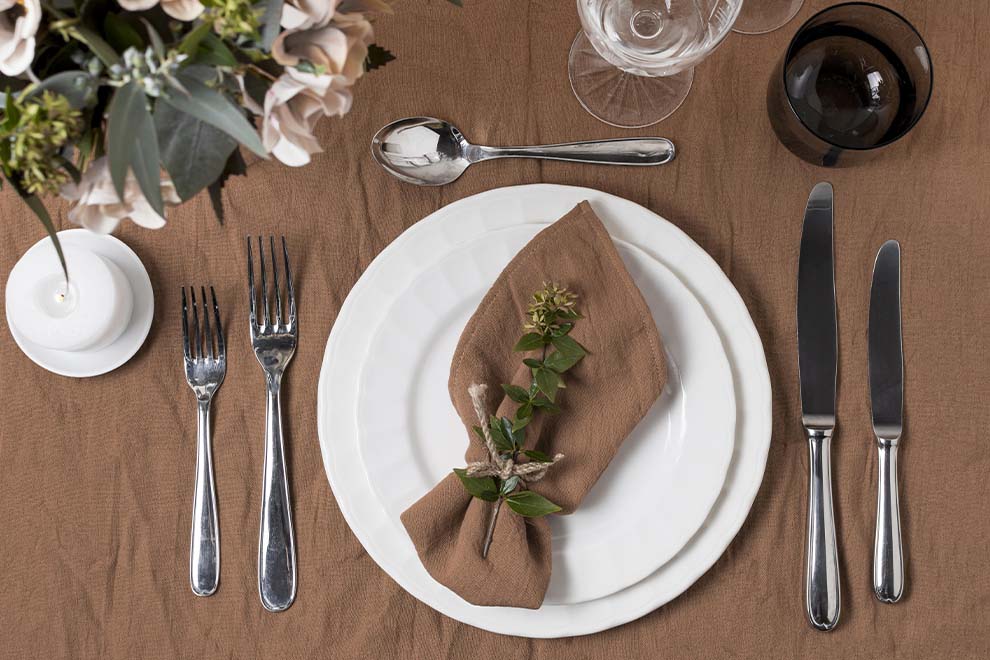 Imagem de uma mesa de madeira montada com pratos, talheres, taças, guardanapo e decorações, vistos de cima. 