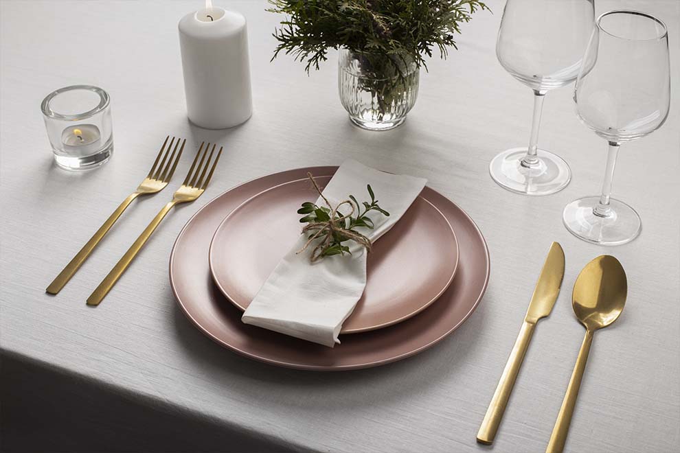 Imagem de uma mesa clara de madeira, montada com um par de pratos, guardanapo, taças, talheres e um vaso de plantas.