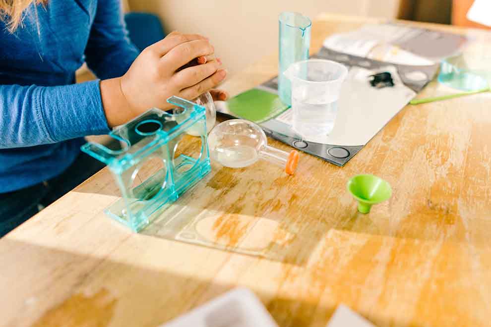 Adolescente brincando com seu kit de ciencia e experimentos em cima de uma mesa de madeira da sala.